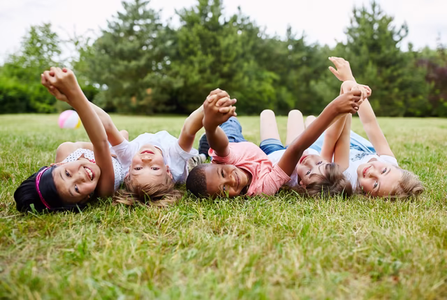 Kinder liegen auf dem Rücken in der Wiese, mit den lachenden Gesichtern zur Kamera gewandt