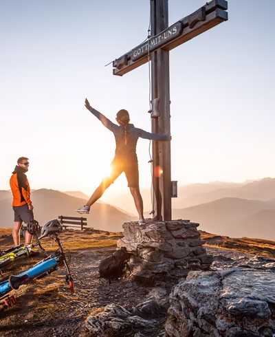 Junge Mountainbikerin am Gipfelkreuz in der Morgensonne, Freund lacht sie an