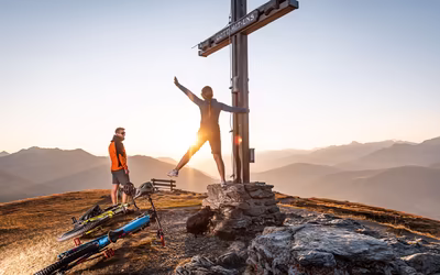Junge Mountainbikerin am Gipfelkreuz in der Morgensonne, Freund lacht sie an