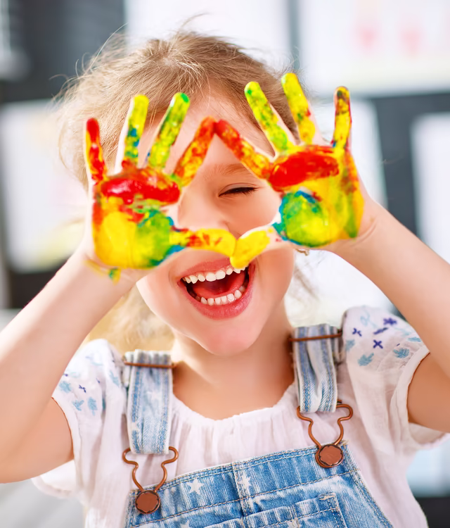 Kind hält mit bunten Farben bemalte Handflächen in die Kamera, grinst