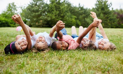 Fröhliche Kinder liegen im Gras mit dem Kopf nach hinten, Blick Richtung Fotograf © Robert Kneschke